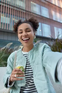 happy-optimistic-woman-with-two-hair-buns-dressed-jacket-enjoys-free-time-walking-city-holds-bottle-detox-drink